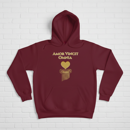 A woman wearing a maroon hoodie with a gold graphic print that reads 'AMOR VINCIT OMNIA' above a gold heart and column design, standing against a white brick wall.