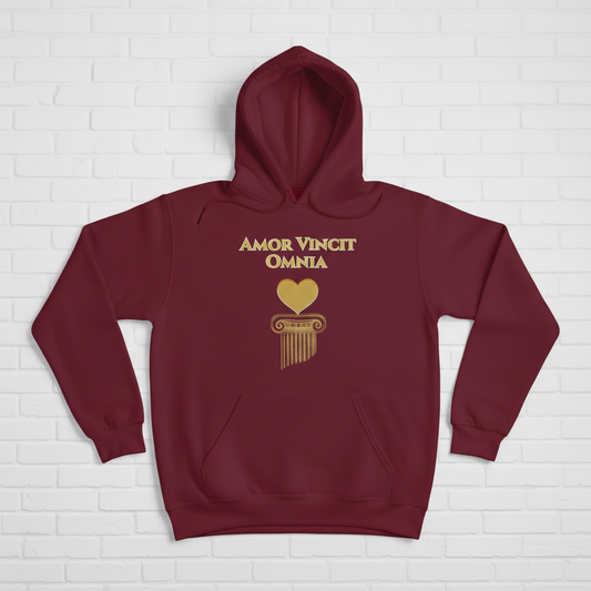 A woman wearing a maroon hoodie with a gold graphic print that reads 'AMOR VINCIT OMNIA' above a gold heart and column design, standing against a white brick wall.
