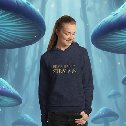 A woman wearing a navy blue hoodie with 'BEAUTIFULLY STRANGE' printed in gold text on the chest, set against a whimsical blue forest background with giant mushrooms.