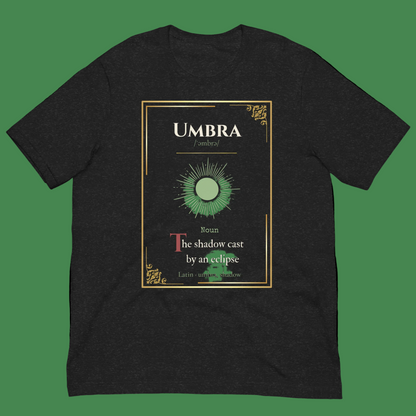 A black heather crewneck t-shirt featuring a graphic print of a dictionary definition card for the word 'UMBRA'. The card has a gold border, a green sun illustration, and text defining umbra as 'the shadow cast by an eclipse'.