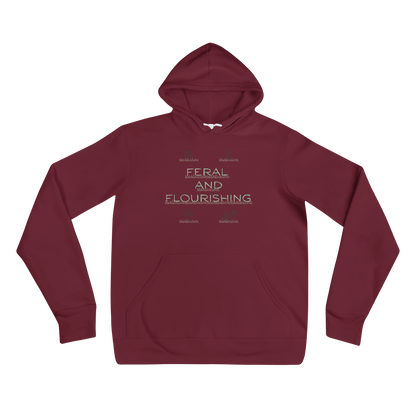 A maroon pullover hoodie with a front kangaroo pocket and a light beige graphic text print reading 'FERAL AND FLOURISHING' on the chest, displayed against a solid white background.